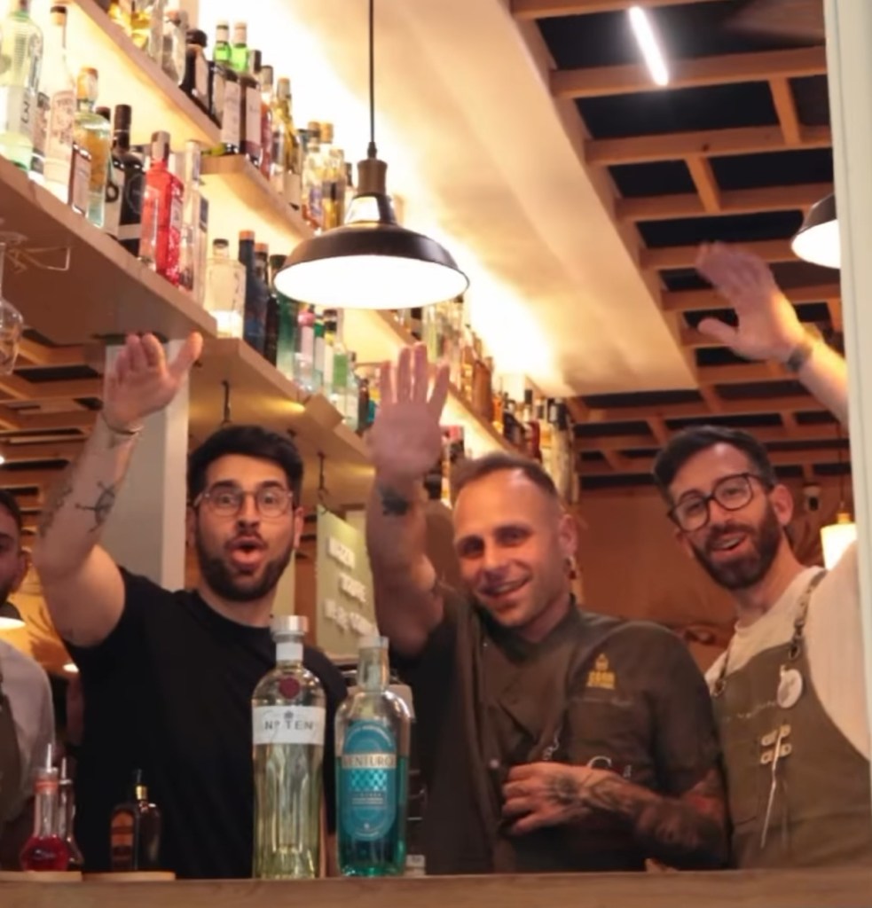 Quattro barman sorridenti con le mani alzate dietro il bancone di un bar, con bottiglie di alcol sullo sfondo.
